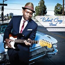 Black man with brown hat plays a brown and yellow guitar standing in front of a light blue car.