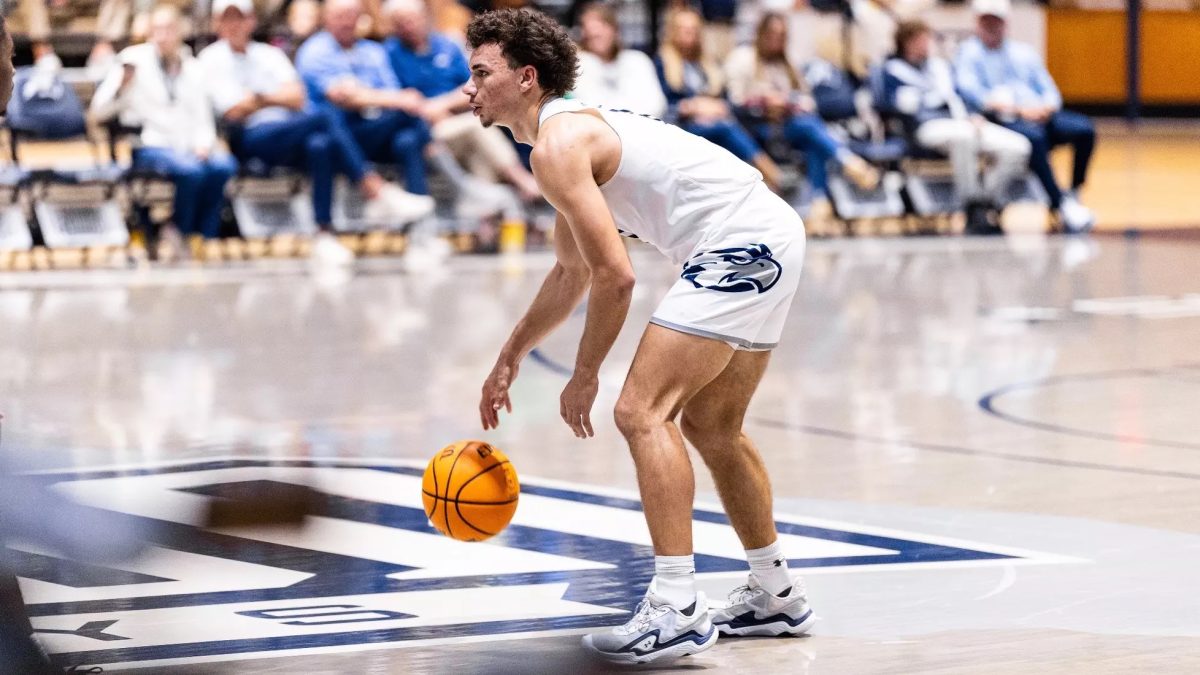 A basketball player wearing a white jersey and shorts dribbles a basketball on a grey court.