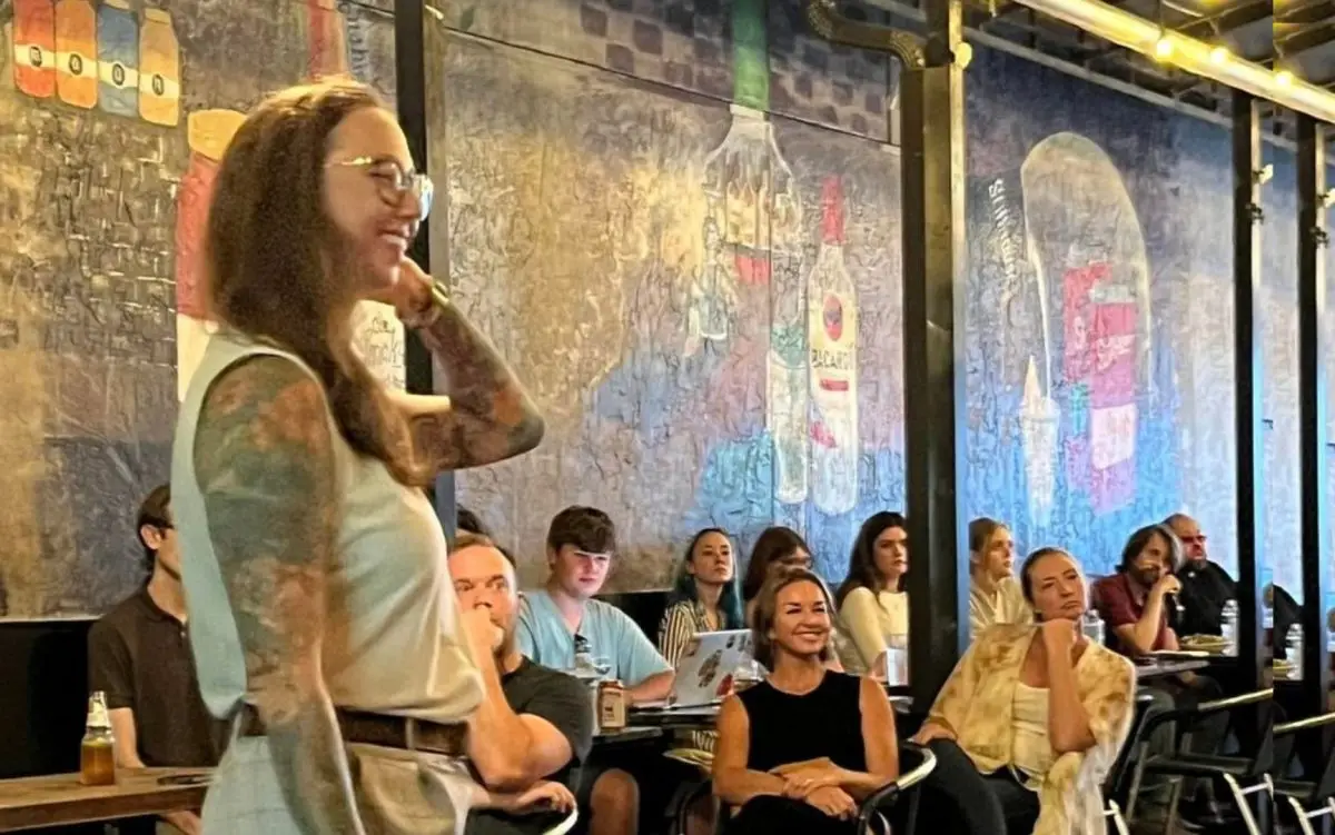 Woman in white tank looks to the right. A crowd sitting behind her looks in same direction.