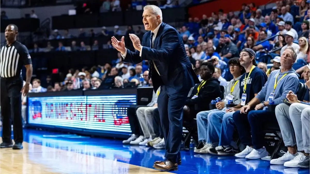 A head coach wearing a navy suit motions his hands in a clapping motion on a blue basketball court.