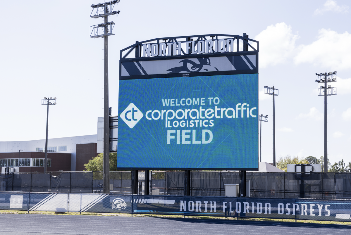 A blue jumbotron above a sports field and track that reads, "Welcome to Corporate Traffic Logistics Field."