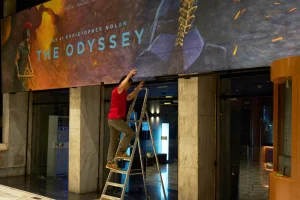 A worker in a red shirt stands on a ladder, installing a billboard that shows an over-the-shoulder shot of a suit of armor next to "The Odyssey" in blue letters