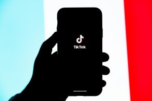 Graphic shows blue, white, and red striped background. A blacked out hand holds a cellphone with a TikTok logo on the screen.